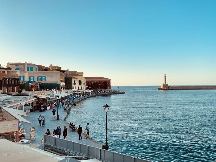Seaside view of a Greek village harbor with people walking along the waterfront and a lighthouse in the distance. Seaside view of a Greek village harbor with people walking along the waterfront and a lighthouse in the distance.