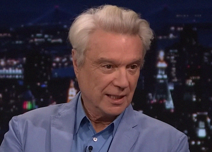 Man with white hair wearing a blue blazer speaking about meeting famous people and sharing celebrity experiences on a nighttime show.