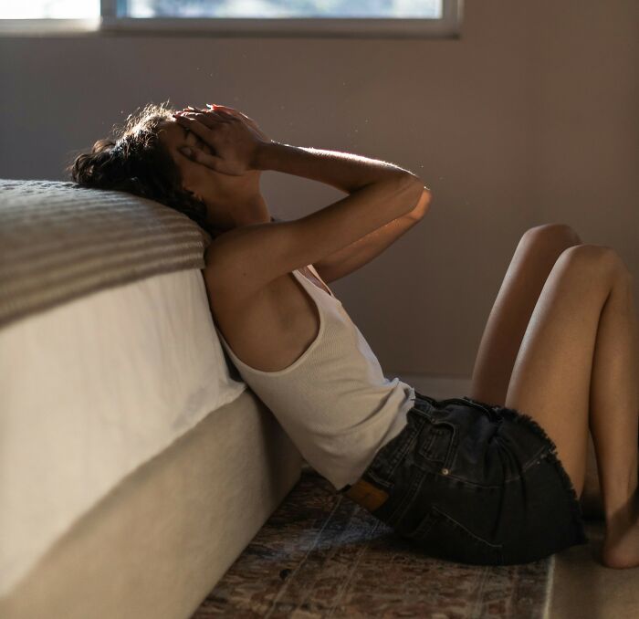 Woman sitting on the floor beside bed, holding head in distress, depicting men being men and women suffering concept.