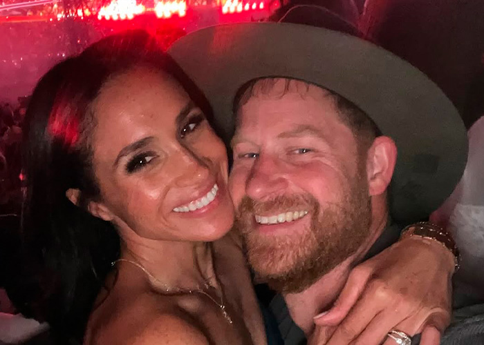 Meghan Markle and Prince Harry smiling closely together at a dimly lit indoor event with red lights. Meghan Markle and Prince Harry smiling closely together at a dimly lit indoor event with red lights.