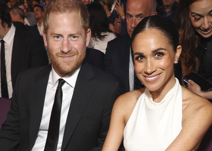 Prince Harry and Meghan Markle smiling at a formal event with attendees seated around them in the background. Prince Harry and Meghan Markle smiling at a formal event with attendees seated around them in the background.