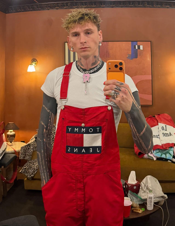 Machine Gun Kelly taking a mirror selfie wearing red overalls and a white shirt with tattooed arms visible.