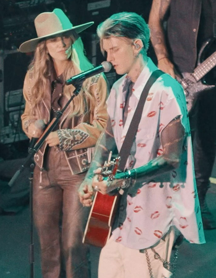 Machine Gun Kelly performing on stage with guitar, accompanied by a female singer wearing a hat and western-style outfit.