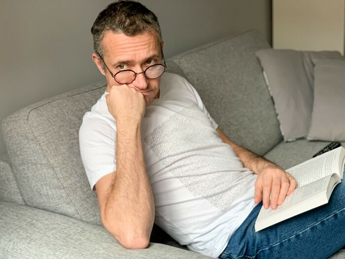 Man with glasses reading a book on a couch, representing incredible minds who proved you don’t need a degree to be a genius.
