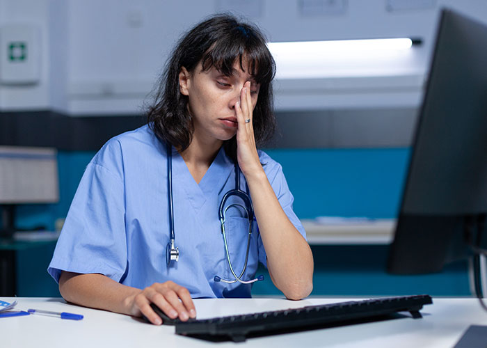Tired woman in medical scrubs with stethoscope at computer, reflecting frustration after reheating pizza leftovers. Tired woman in medical scrubs with stethoscope at computer, reflecting frustration after reheating pizza leftovers.