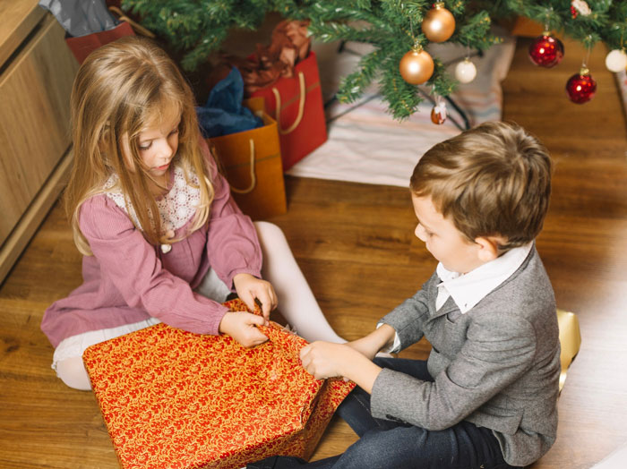 Two children sitting by a Christmas tree, unwrapping a large festive gift during holiday celebrations. Two children sitting by a Christmas tree, unwrapping a large festive gift during holiday celebrations.