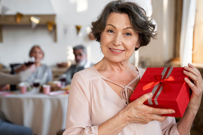 Older woman smiling and holding a red gift box during a family Christmas gathering at home. Older woman smiling and holding a red gift box during a family Christmas gathering at home.