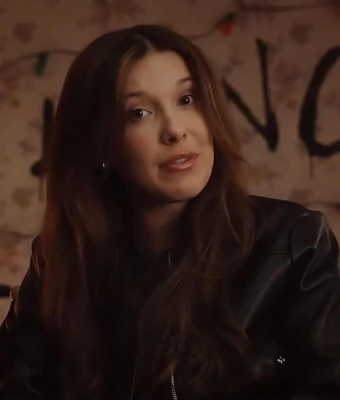 Millie Bobby Brown in a leather jacket sharing a sad confession about plastic surgery rumors in a dimly lit setting.