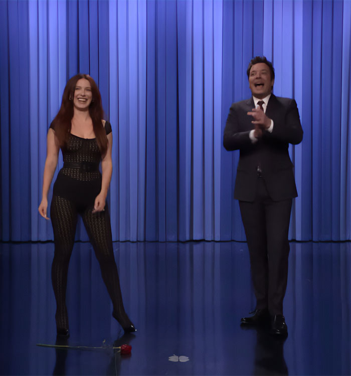 Actress in a sheer black outfit standing next to a host on a stage with blue curtains during a Fallon appearance. Actress in a sheer black outfit standing next to a host on a stage with blue curtains during a Fallon appearance.