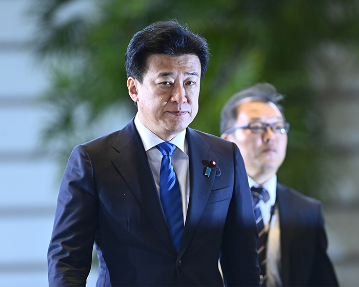 Japanese government official in a dark suit and blue tie walking with another man amid greenery background during a formal event Japanese government official in a dark suit and blue tie walking with another man amid greenery background during a formal event