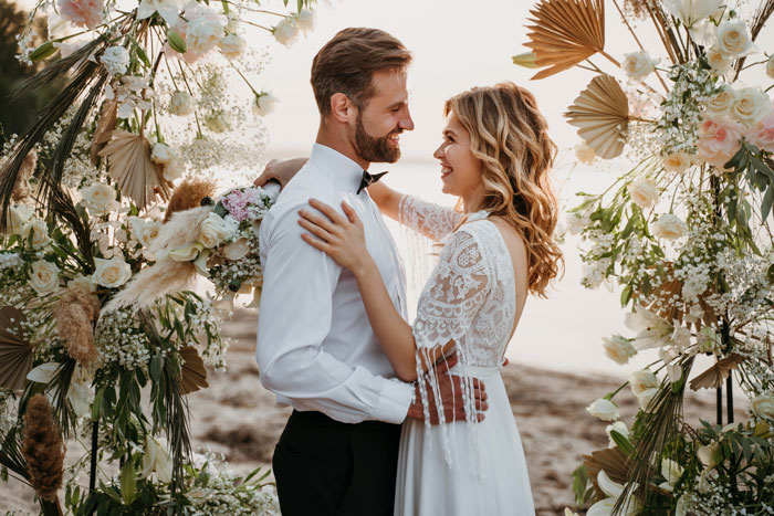 Bride and groom smiling at each other during outdoor wedding surrounded by floral decorations and greenery.