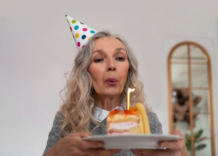 Older woman wearing a party hat blowing out a birthday candle, illustrating mom rages at son for forgetting birthday. Older woman wearing a party hat blowing out a birthday candle, illustrating mom rages at son for forgetting birthday.