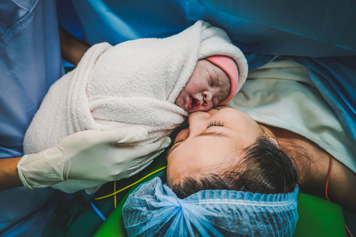 Newborn baby wrapped in a blanket held by medical staff while mother wearing surgical cap lies in a hospital bed after labor. Newborn baby wrapped in a blanket held by medical staff while mother wearing surgical cap lies in a hospital bed after labor.