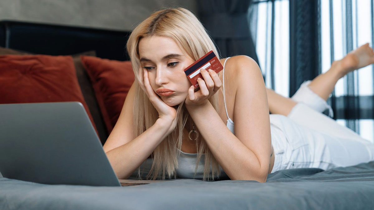 Young woman laying on bed looking at laptop in disbelief holding credit card, stressed about identity theft and ruined credit.