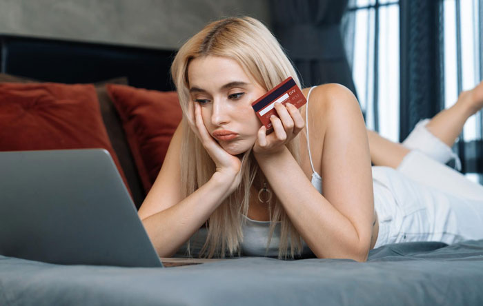 Young woman holding credit card looking worried at laptop screen illustrating mom nuked credit score concerns online.