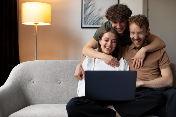 Young woman and two men smiling while using a laptop on a couch, illustrating family dynamics and relationship challenges. Young woman and two men smiling while using a laptop on a couch, illustrating family dynamics and relationship challenges.