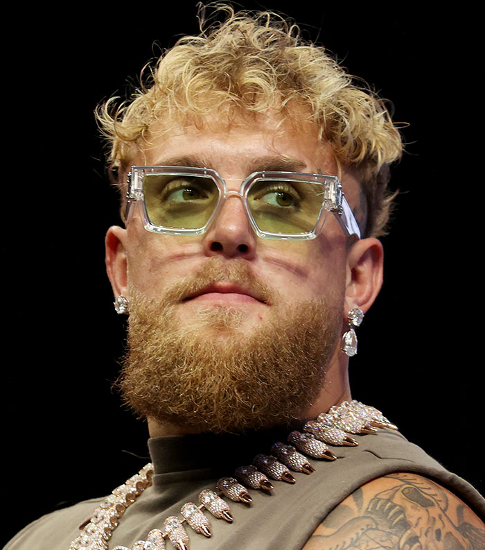 Bearded man with curly blonde hair wearing green tinted glasses and diamond jewelry representing most disliked celebrities 2025.