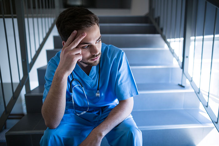 Young male doctor in blue scrubs sitting on stairs, looking thoughtful about wrong self-diagnoses in a medical setting.