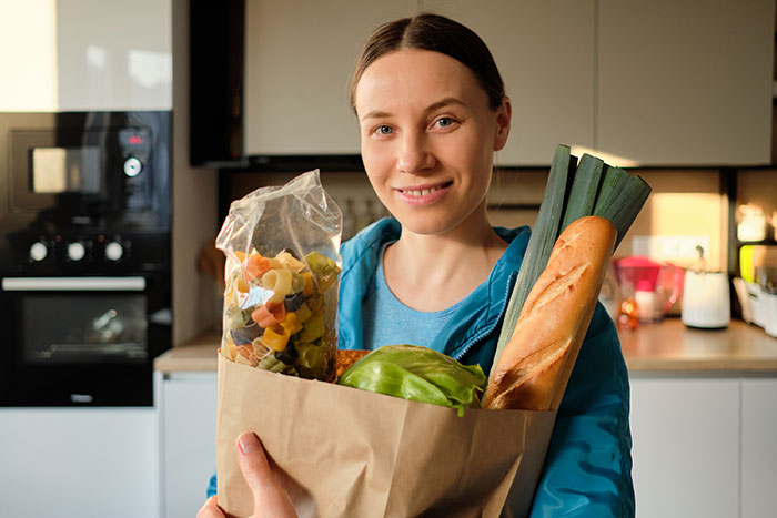 Woman holding groceries in kitchen, representing mom accused of embarrassing son’s girlfriend who offered to cook meals. Woman holding groceries in kitchen, representing mom accused of embarrassing son’s girlfriend who offered to cook meals.