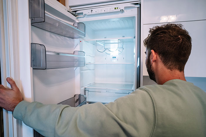 Man in green sweater looking into an empty fridge after hosting an expensive Thanksgiving dinner, surprised no leftovers remain.