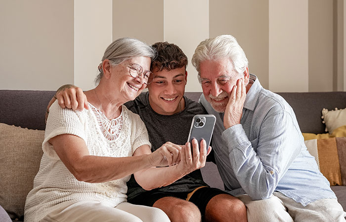Exhausted teen smiling with grandparents on couch, all looking at smartphone.