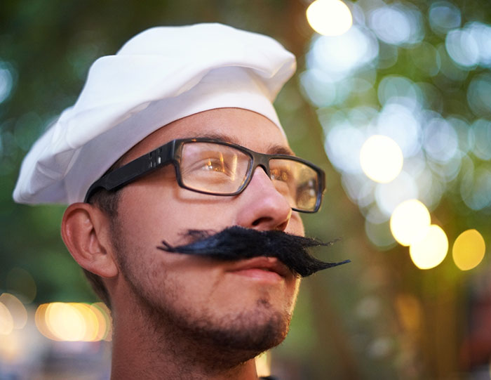 Man from different countries wearing a white chef hat and fake mustache, denying popular myths foreigners believe outdoors.