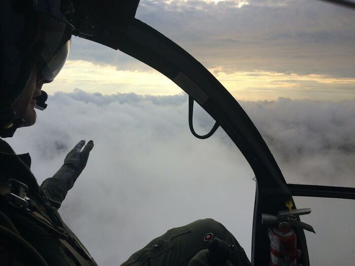 Pilot in helicopter cockpit flying above clouds during sunrise, showcasing unique jobs that make a 9-to-5 more bearable.