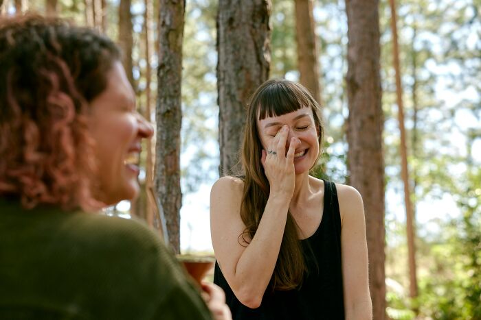 Two women laughing outdoors among trees, illustrating harmless but toxic habits in casual social interaction.