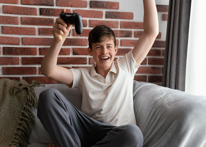 Teen boy playing video games on couch, celebrating a win, showing entitlement after using credit card for games.