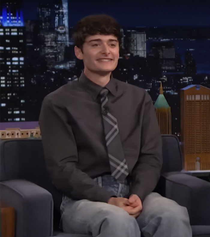Noah Schnapp sitting on a talk show set, wearing a dark shirt and plaid tie, amid Stranger Things season acting discussion.