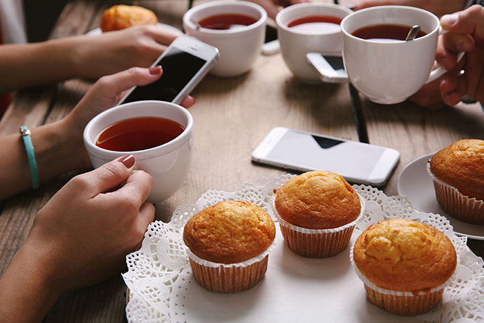 People holding cups of tea at an office potluck with store bought desserts and smartphones on the wooden table. People holding cups of tea at an office potluck with store bought desserts and smartphones on the wooden table.