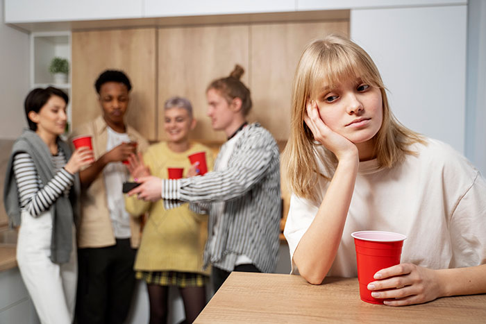 A young woman feeling left out at an office potluck, holding a red cup while others socialize in the background. A young woman feeling left out at an office potluck, holding a red cup while others socialize in the background.