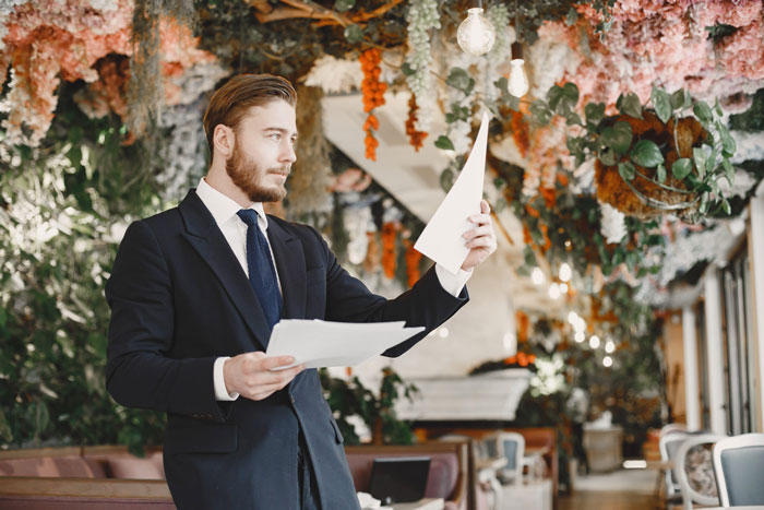Officiant in suit reviewing papers indoors surrounded by floral wedding decorations and hanging greenery.