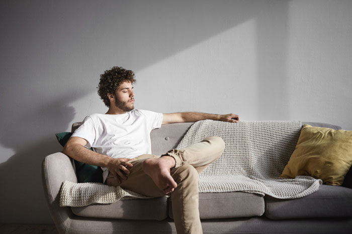 Young man sitting thoughtfully on a gray sofa in a sunlit room, wearing a white t-shirt and beige pants. Young man sitting thoughtfully on a gray sofa in a sunlit room, wearing a white t-shirt and beige pants.