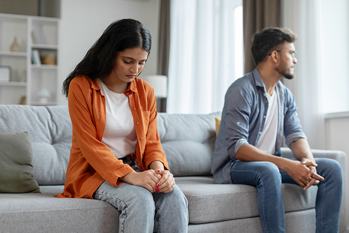 Couple sitting apart on couch, tense after truth about age deception is revealed in relationship discussion. Couple sitting apart on couch, tense after truth about age deception is revealed in relationship discussion.