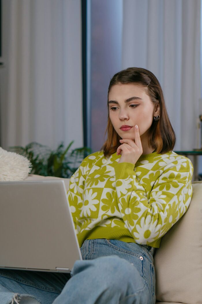 Young woman in floral sweater sitting on couch, thoughtfully using laptop, reflecting on things learned about women in relationships.