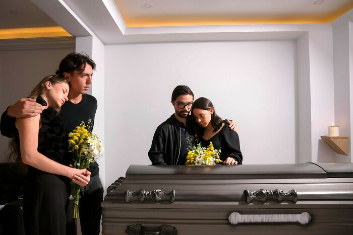 Two grieving couples standing by a casket holding flowers, representing the aftermath of biggest scandals in workplaces.