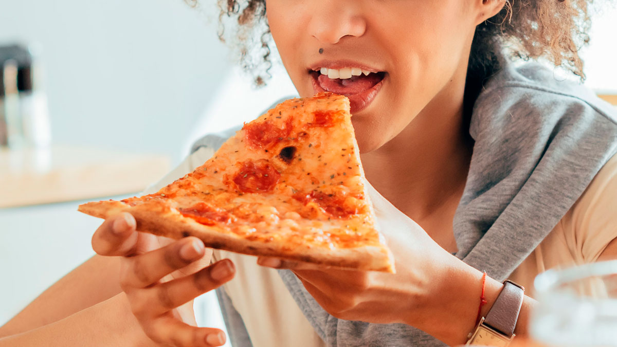Young woman babysitter holding and eating a slice of pepperoni pizza, enjoying a casual moment indoors.