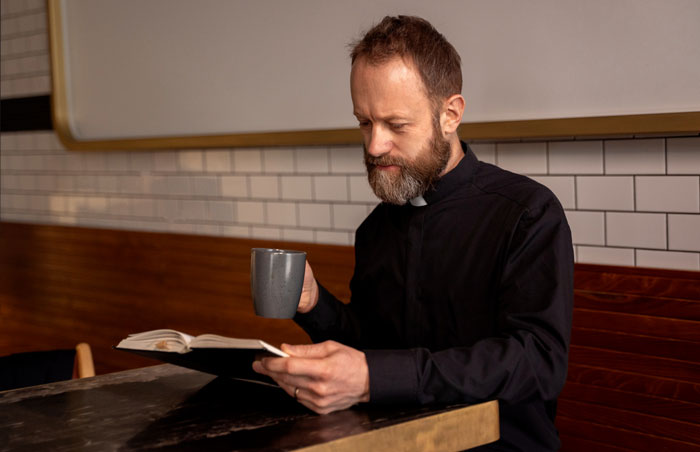 Pastor in black clerical shirt reading a book and holding a mug in a church setting with wooden and tiled walls. Pastor in black clerical shirt reading a book and holding a mug in a church setting with wooden and tiled walls.
