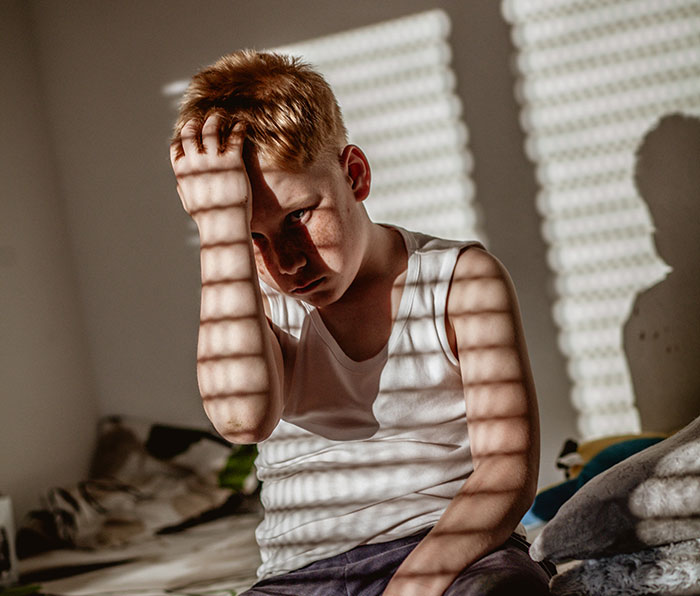 Sad boy with hand on head sitting on bed, shadows from blinds highlighting a moment of selfish unloving sociopath behavior.
