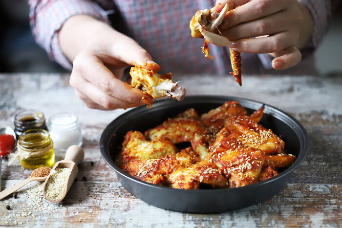 Person eating spicy chicken wings from a black bowl with seasonings on a rustic wooden table.