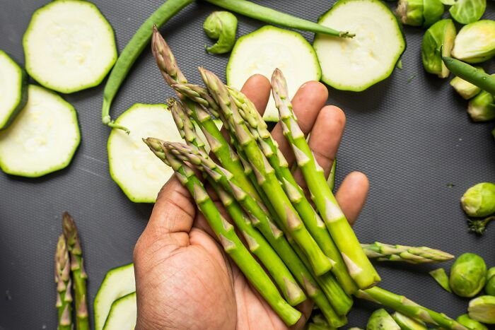 Hand holding fresh asparagus with sliced zucchini and Brussels sprouts on a dark surface, highlighting partner terrors concept.