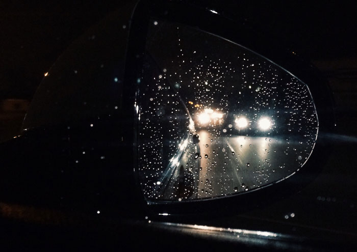 Rain-covered car side mirror reflecting bright headlights at night, evoking a sense of scary moments and trauma.