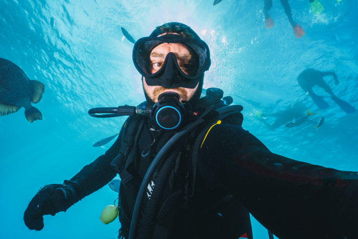 Scuba diver underwater surrounded by fish, capturing a selfie during one of the scariest moments shared by people.