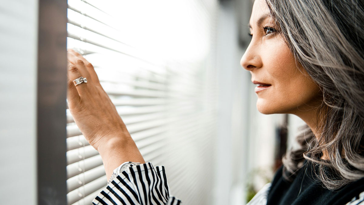 Middle-aged woman with gray hair looking outside through blinds, illustrating snobbish behavior in online shaming context.