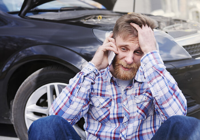Man in plaid shirt looking stressed while talking on phone near a broken-down car, a scary situation people still talk about.