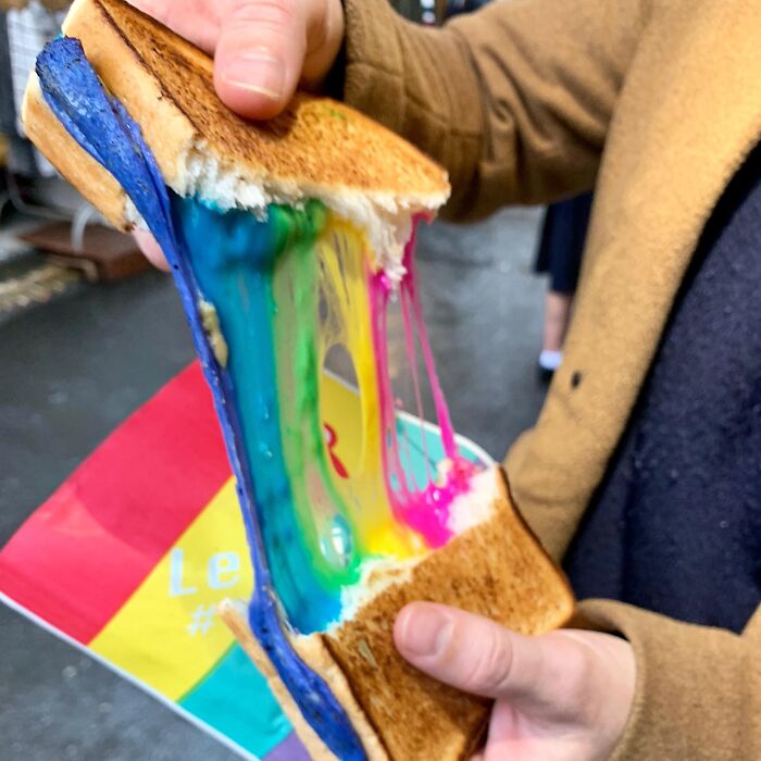 Colorful melted cheese pulls apart from a toasted sandwich held by a person, showing a unique foodie dish.
