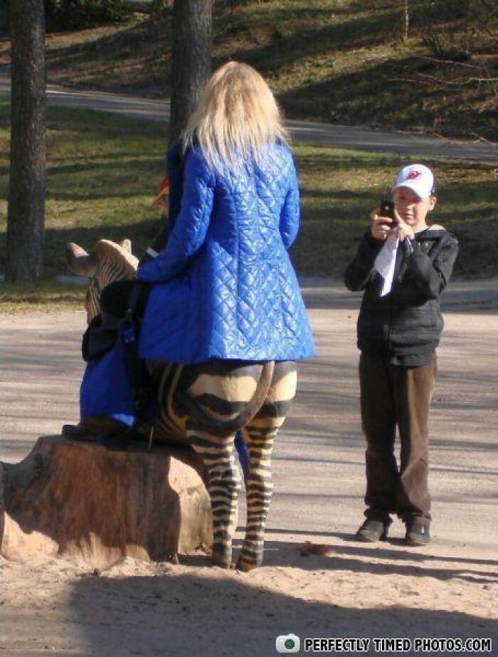 Woman in a blue coat sitting on a zebra statue, with a person taking a photo, showcasing perfectly timed photos.