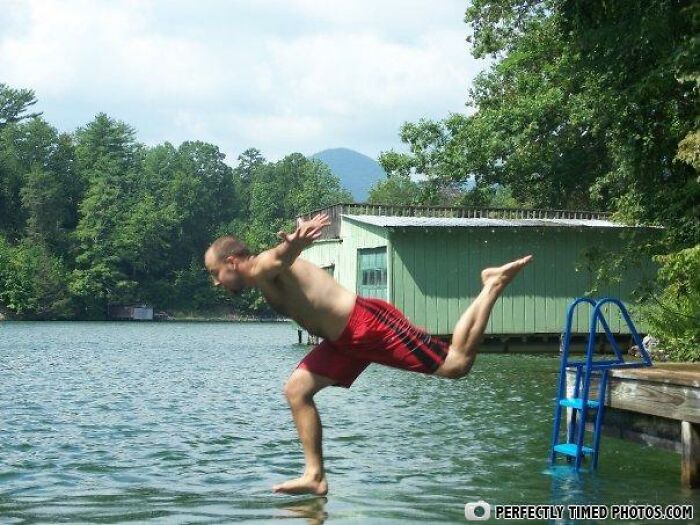 Man in red shorts captured in a perfectly timed photo as he leaps off a dock into a lake surrounded by trees.