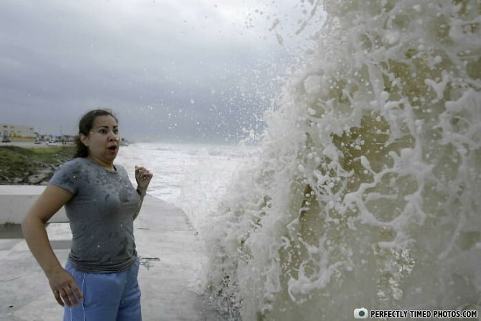 Woman in a gray shirt caught in a perfectly timed photo as a large wave splashes near her on a cloudy day.
